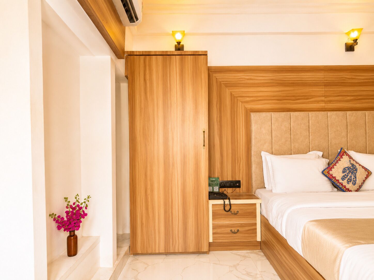 A hotel room with a wooden wardrobe, a bed with white bedding, a bedside table with a phone, and a vase with flowers.