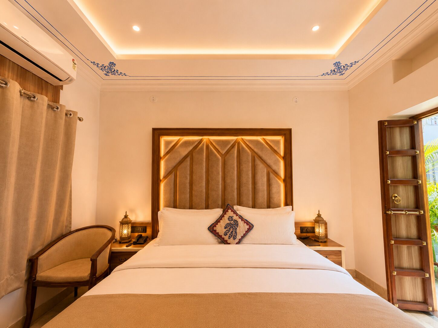 A hotel room with a large bed, a wooden headboard, two bedside tables with lamps, a chair, and a view of greenery outside.