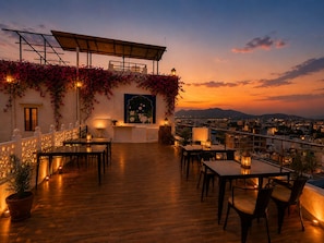 Terraço no telhado com piso de madeira, mesas, cadeiras, plantas em vasos e vista do horizonte urbano ao pôr do sol.