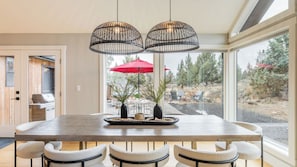 A dining area with a large table, four chairs, two hanging pendant lights, and a view of the outdoors through large windows.