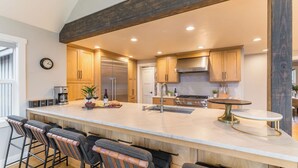 A modern kitchen with wooden cabinets, a stainless steel refrigerator, a built-in oven, and a large island with bar stools. - The Gem Estate by Avantstay Modern Farmhouse w/ Lavender Fields + Hot Tub (Bend)
