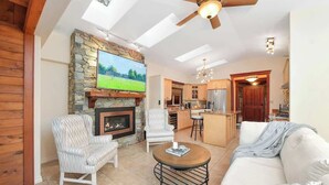 A spacious living room with a fireplace, a television mounted on a stone wall, a ceiling fan, and a kitchen area in the background. - Dolphin House Oceanview Family Getaway (Nanoose Bay)