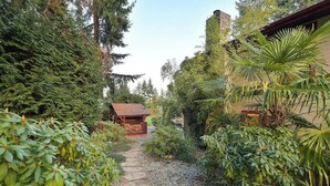 A garden pathway leading to a rustic wooden structure surrounded by lush greenery and various plants. - Dolphin House Oceanview Family Getaway (Nanoose Bay)