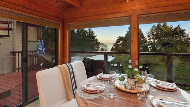 A wooden deck with a dining table set for four, overlooking a lush landscape. - Dolphin House Oceanview Family Getaway (Nanoose Bay)