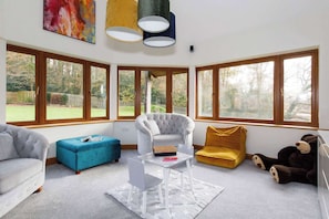 A room with a grey sofa, a blue ottoman, a white armchair, a yellow chair, and a large teddy bear. There are three windows with wooden frames, a white table with a box, and a grey rug with star patterns. - Serene Riverside: 6BR Luxury Retreat in Curdridge (Curdridge)