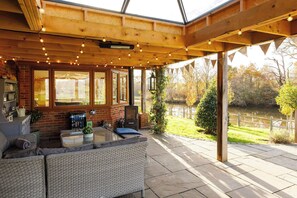 A covered patio area with wicker furniture, a brick fireplace, and a view of a lake. - Serene Riverside: 6BR Luxury Retreat in Curdridge (Curdridge)