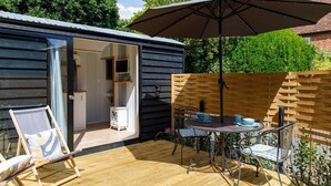 Una terraza de madera con mesa y sillas, un puerta corrediza de vidrio que da a una habitación y una sombrilla grande para dar sombra.