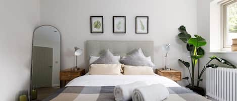 A modern bedroom with a large bed, a headboard, bedside tables, a mirror, and framed botanical prints on the wall.