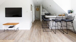 A modern kitchen with a dining area, featuring a TV mounted on the wall, a wooden table, and black wireframe chairs. - Stylish Self-Contained Countryside Accommodation (Funtington)