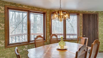 A dining room with a wooden table, six chairs, a chandelier, and large windows with a view of trees.