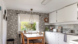 Et køkken med hvide skabe, et træspisebord med stole, en hængende lampe og et vindue med udsigt til grøn natur.