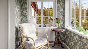 Et hyggeligt værelse med en fletstol, et lille bord med en vase blomster og et vindue med udsigt til træer.