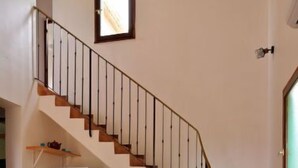 A compact living space with a staircase, a dining area with chairs and a table, a sofa with a patterned cushion, and a small kitchen area with a refrigerator.