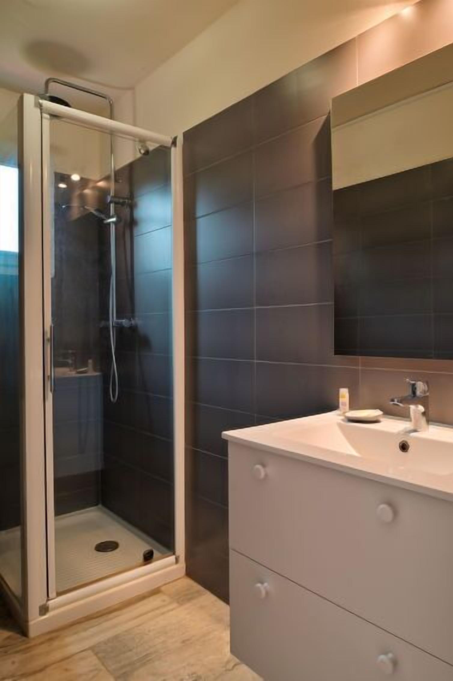 Un baño moderno con cabina de ducha de vidrio, paredes cubiertas de baldosas oscuras y un mueble de tocador blanco con cajones.