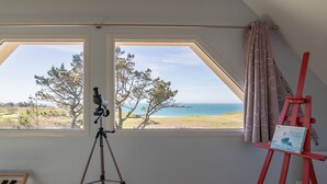 Quarto com grande janela com vista para paisagem costeira, um tripé e um cavalete vermelho com uma pintura.