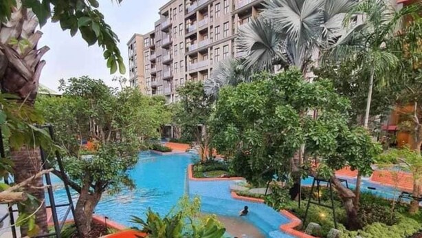 A swimming pool with a slide, surrounded by lush greenery and a multi-story building in the background.