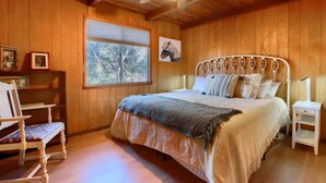 Une chambre aux murs lambrés, avec un lit, une chaise, une table de chevet et une fenêtre équipée de stores.