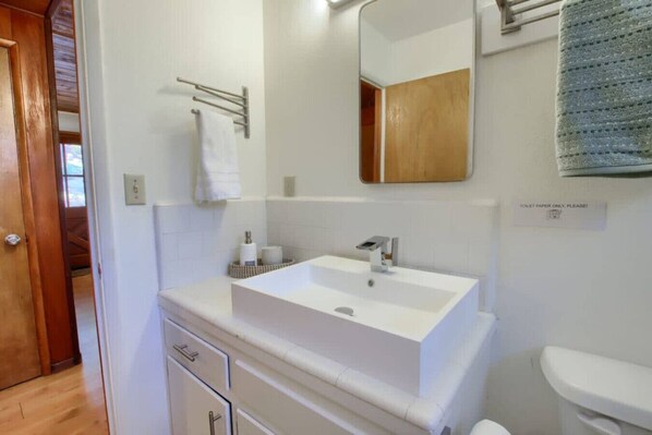Une salle de bain avec un lavabo blanc, un miroir et des toilettes. On y trouve un porte-serviettes avec une serviette blanche et un panneau indiquant : « Veuillez utiliser uniquement le papier toilette ! »