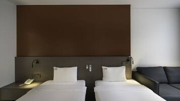 A hotel room with two single beds, a sofa, a bedside table with a phone, and a wall-mounted control panel.