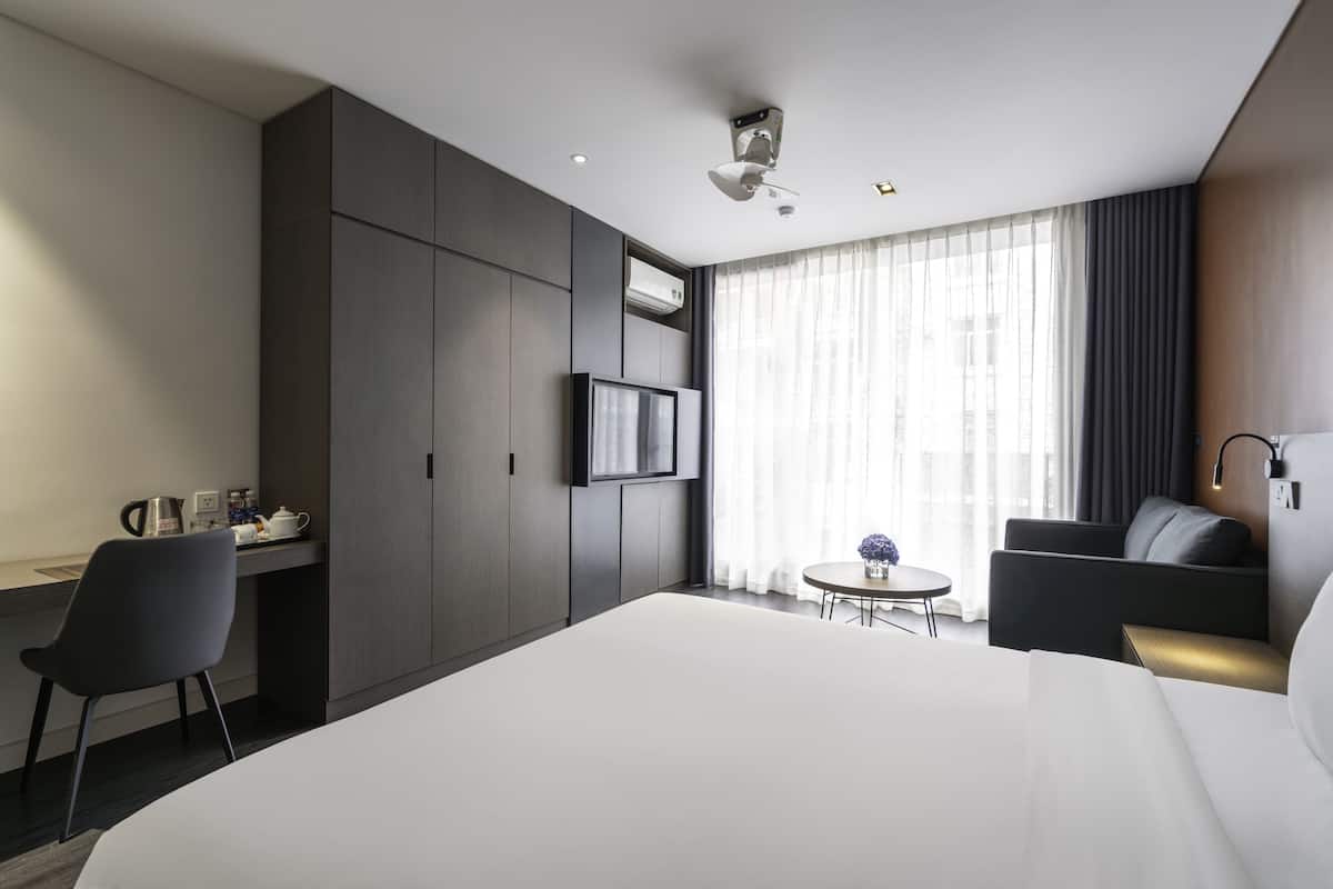 Quarto de hotel moderno com uma cama grande, uma escrivaninha com cadeira, uma mesinha redonda com um vaso e um sofá.