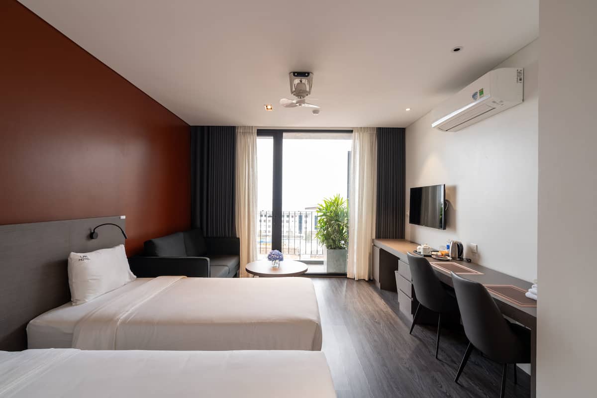 Quarto de hotel com duas camas, uma escrivaninha, uma cadeira, uma televisão e uma varanda com planta.