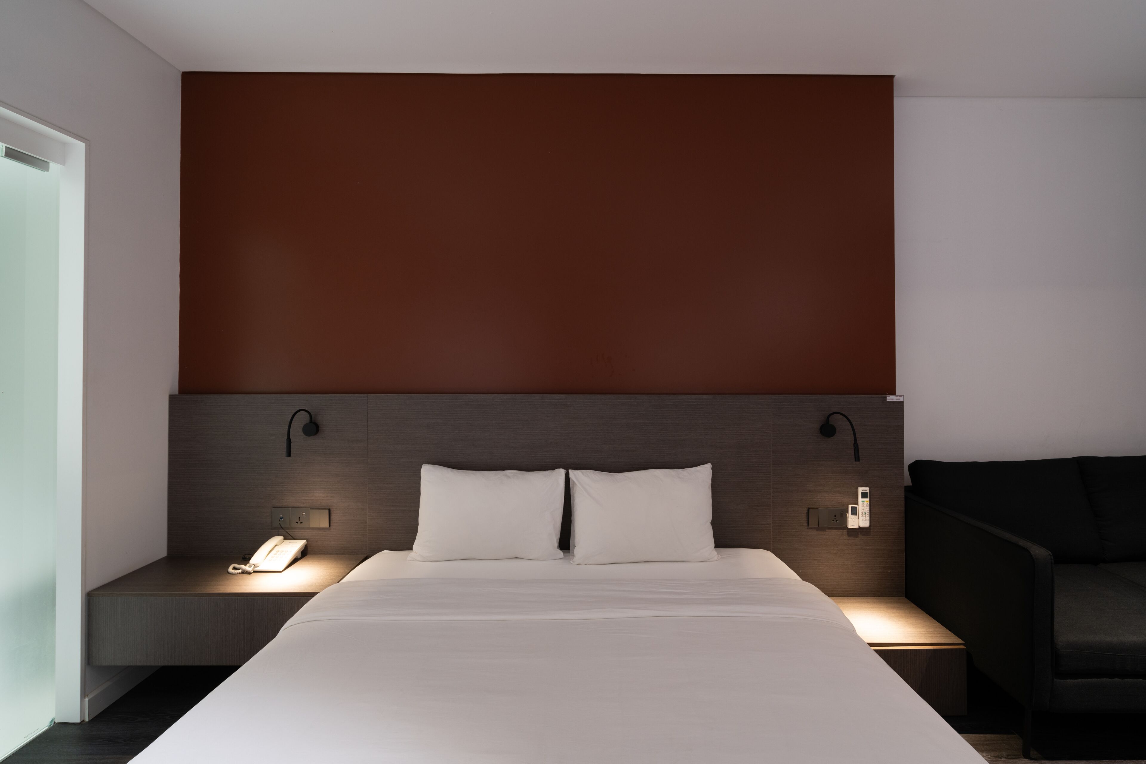 Quarto de hotel moderno com uma cama grande, mesas de cabeceira, telefone e luminária fixada na parede.