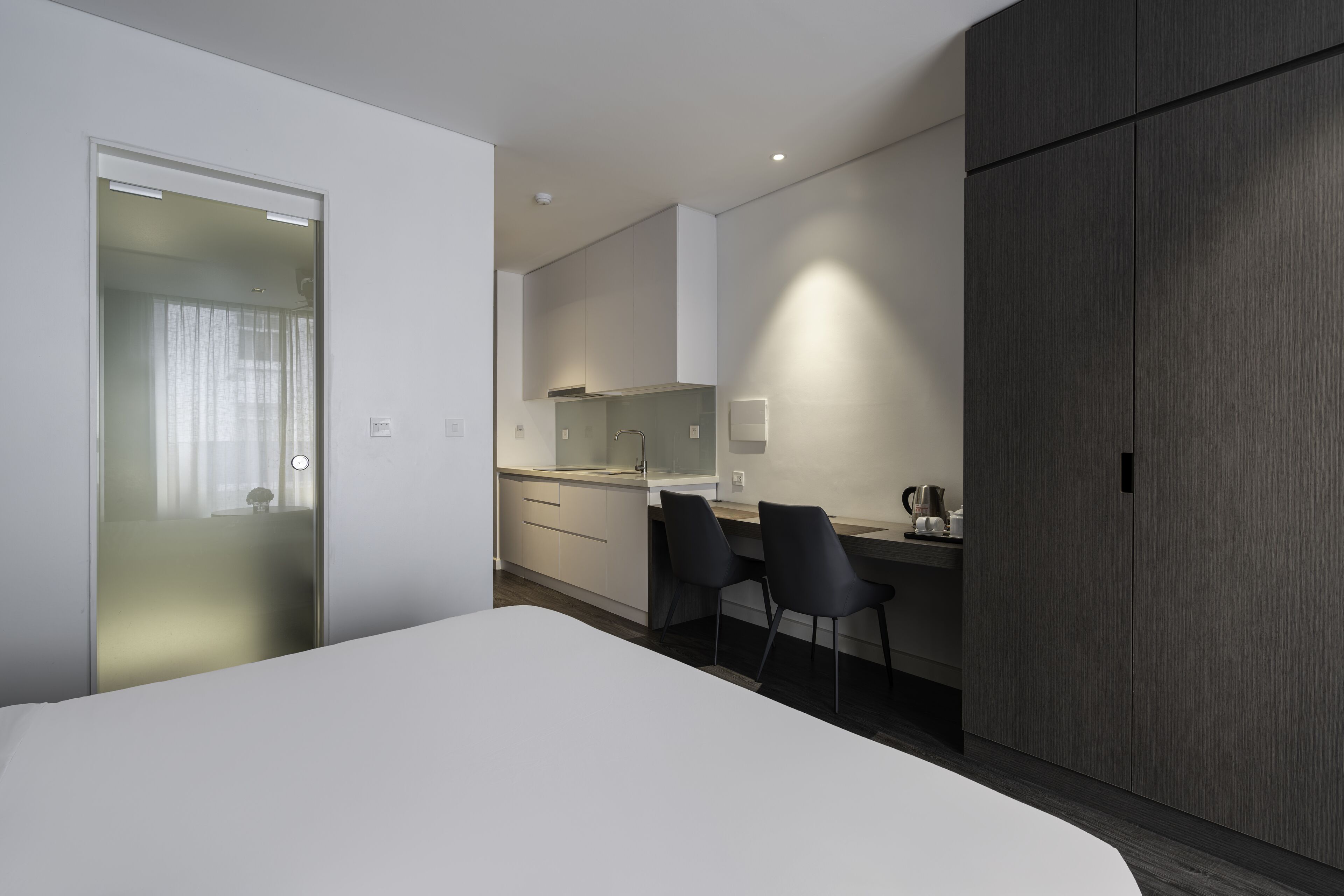 Habitació d'hotel moderna amb un llit, una taula de treball amb dues cadires, una cuineta i un armari.