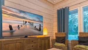 Una habitación acogedora con una tele grande sobre un mueble de madera, sillas de mimbre, una mesa y vistas al exterior a través de una ventana.
