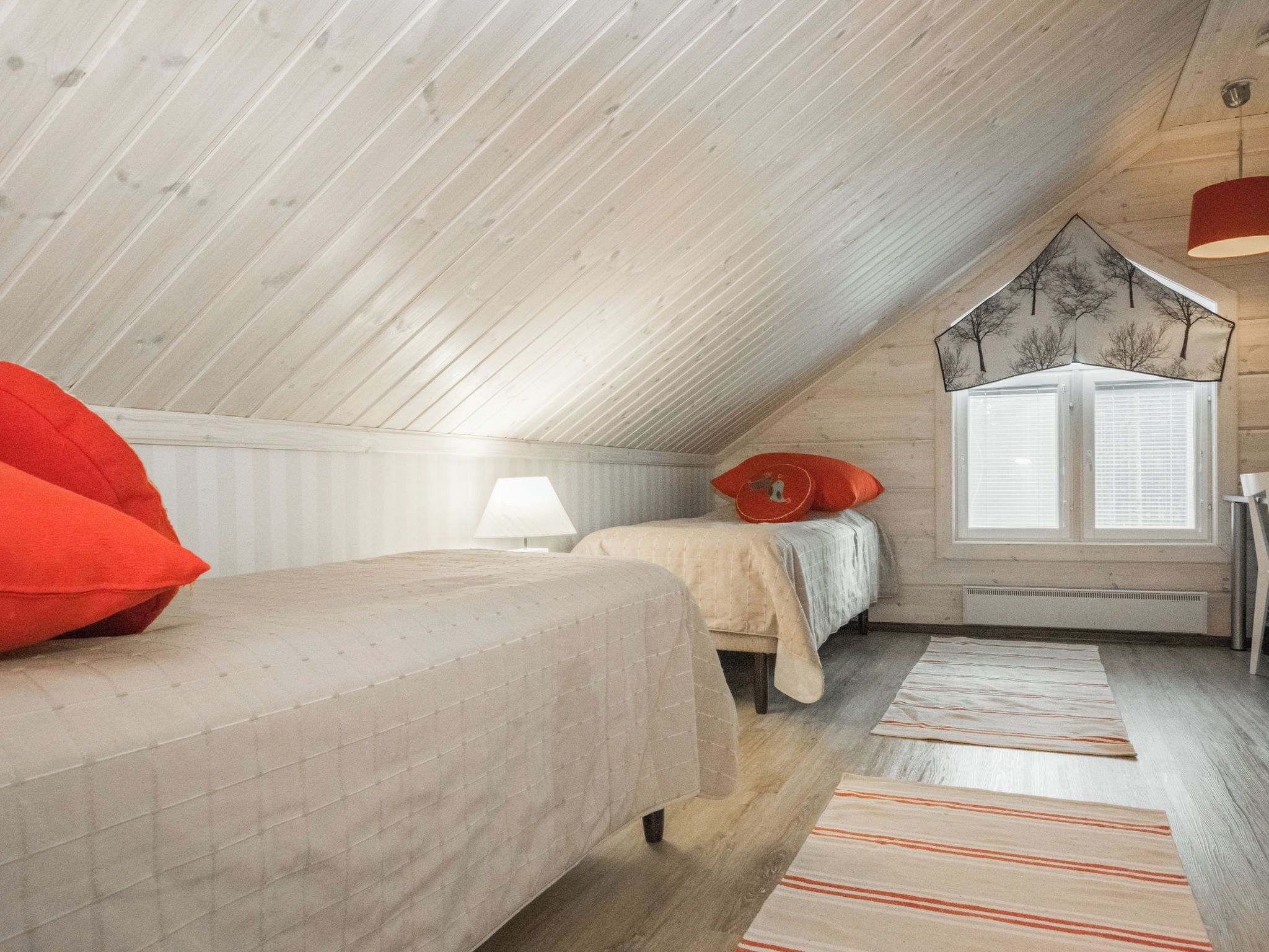 A cozy attic bedroom with a sloped ceiling, two beds with red and white bedding, a window with blinds, and a hanging lamp.