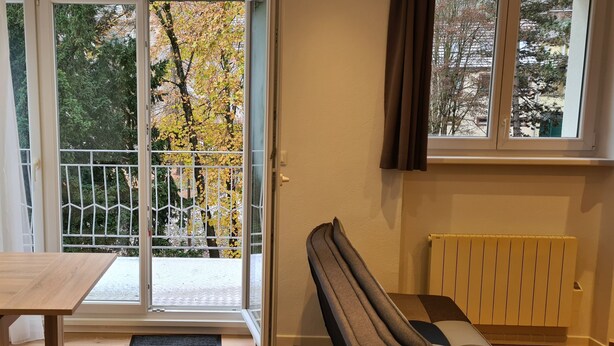 A room with a wooden table, two water bottles, a chair, a radiator, and a view of trees through a window. - Meublés du 25 N°5 à Niederbronn-les-bains (Niederbronn-les-Bains)