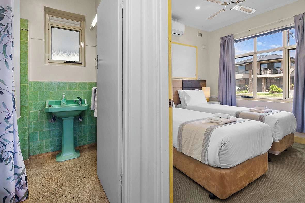 Ein Hotelzimmer mit grünen Fliesen, einem Waschbecken, zwei Betten, einem Fenster mit Blick und einem Deckenventilator.