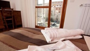 Una camera d'albergo con un letto, una finestra che si affaccia su degli edifici e una sedia di legno.