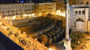 Un antico anfiteatro con una statua centrale, circondato da architettura classica e una piazza ben illuminata.