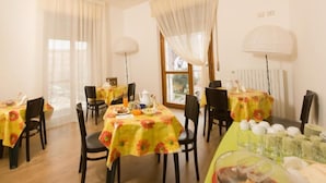 Zona pranzo con tavoli rotondi rivestiti da tovaglie floreali gialle, sedie nere e un tavolo buffet con cibo e bevande.