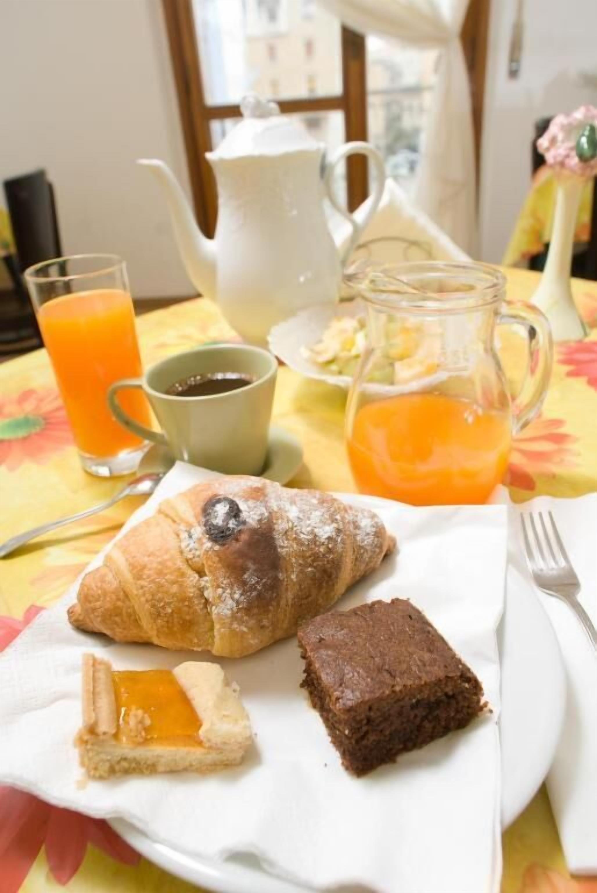 Un desayuno con croissants, una porción de pastel, un brownie en un plato, además de un vaso de jugo de naranja y una taza de café.