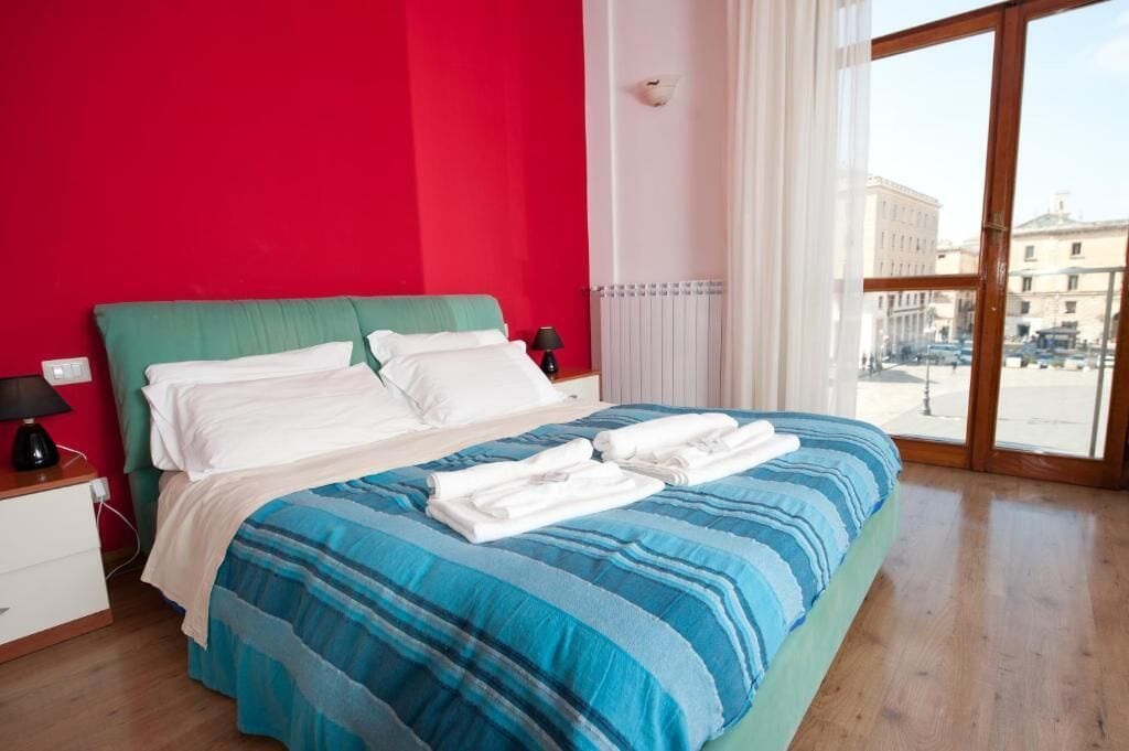 A hotel room with a large bed, a red wall, and a view of the city through a large window.