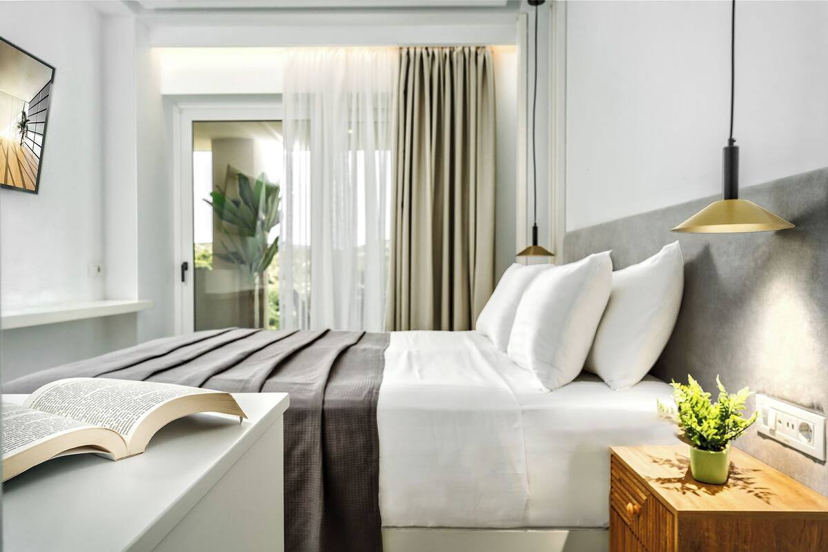 Un dormitorio moderno con cama, mesita de noche, lámpara y vistas al exterior.