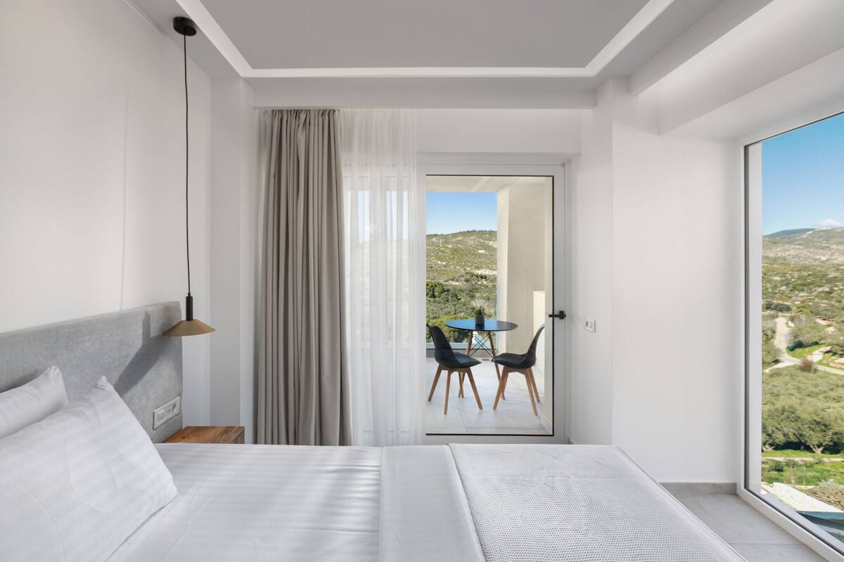 Habitación de hotel moderna con una cama grande, una mesita de noche, una lámpara y vistas a un balcón con sillas y una mesa.
