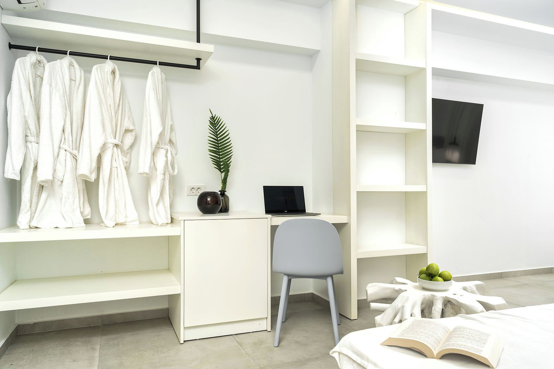 A modern, minimalist bedroom with a bed, a desk with a laptop, a chair, and a shelf with robes.