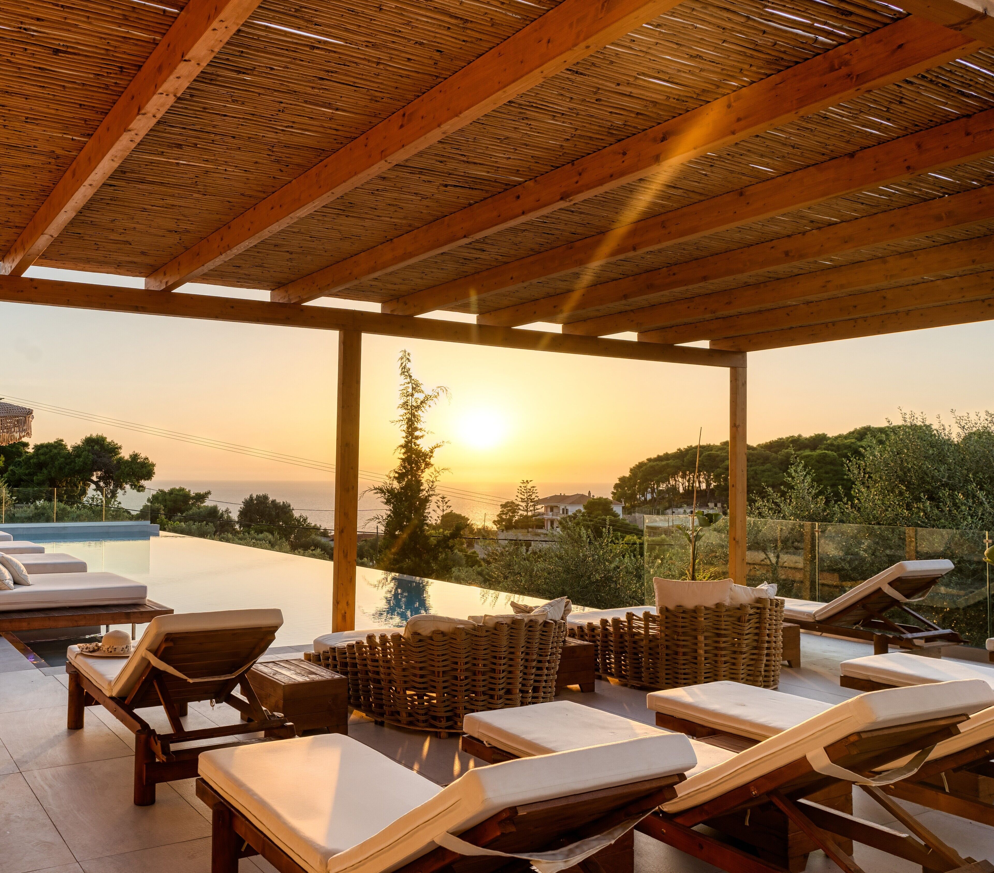 A sunlit outdoor lounge area with wooden beams, lounge chairs, and a view of the sunset over a body of water.