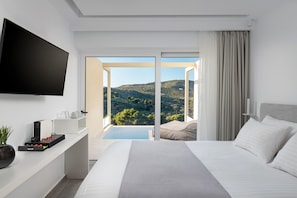 A modern hotel room with a large bed, a flat-screen TV, a coffee machine, and a view of a mountainous landscape through a sliding glass door. - Myzith Luxury Suites  (Zakynthos)
