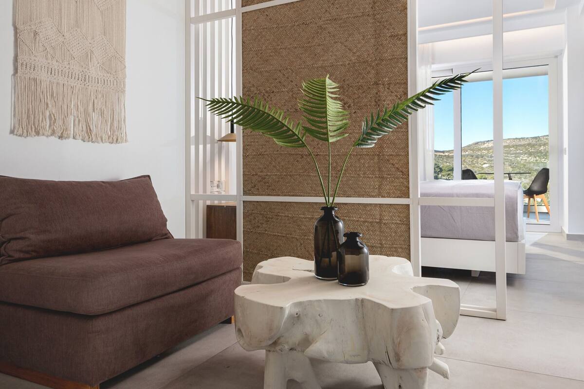 Una sala de estar moderna con un sofá marrón, una mesa de centro blanca con una planta y un amplio ventanal con vistas al exterior.