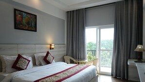 A hotel room with a large bed, a painting on the wall, a balcony with a chair, and a table with a lamp. - AAR Resort and Spa (Dehradun)