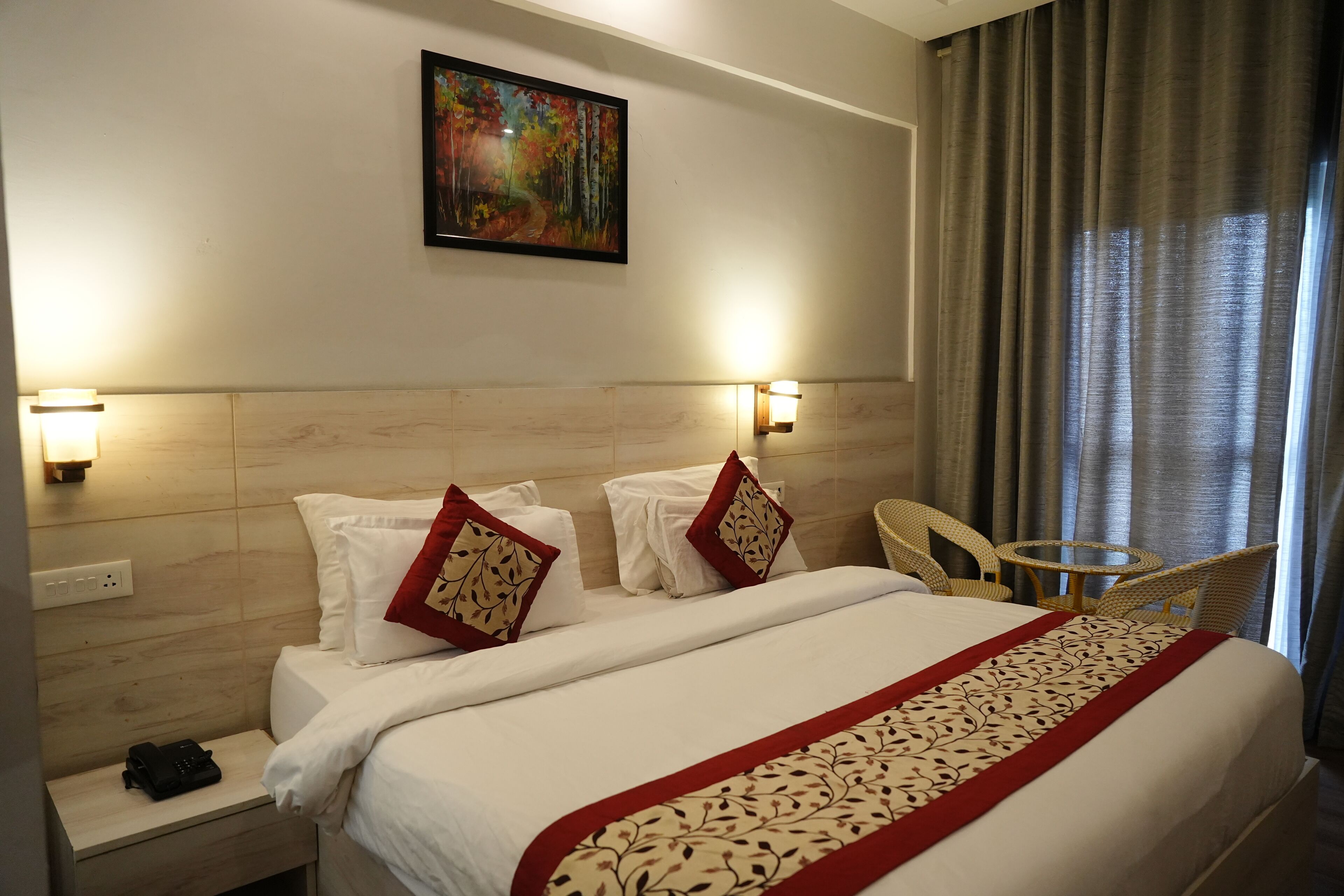 A hotel room with a large bed, two bedside tables, a small round table, and a chair. There is a painting on the wall and a window with curtains.