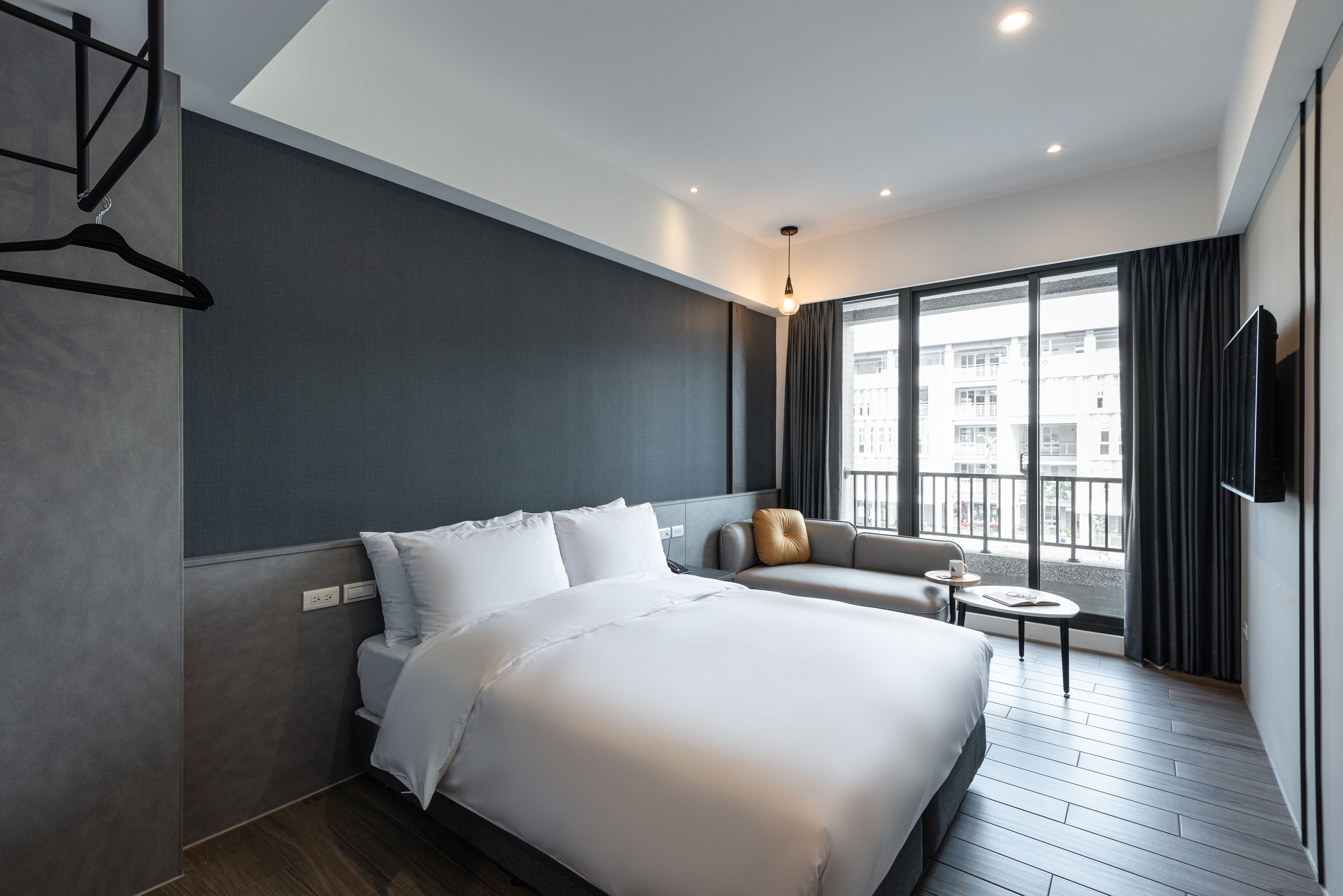 A modern hotel room with a large bed, a sofa, a small table, and a view of the city through large windows.
