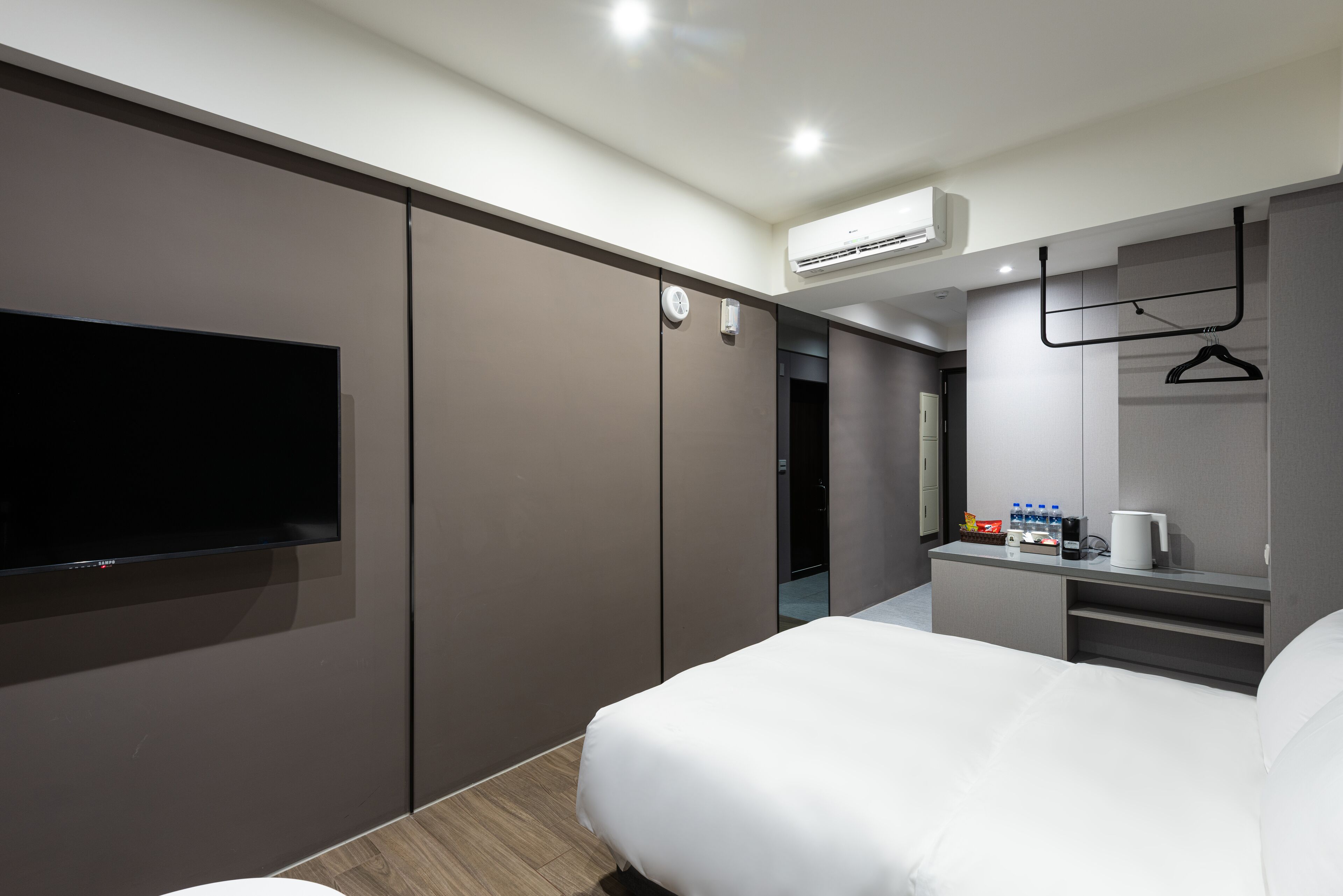 A modern hotel room with a large bed, a flat-screen TV mounted on the wall, a small desk with a coffee maker, and a wardrobe with a mirror.