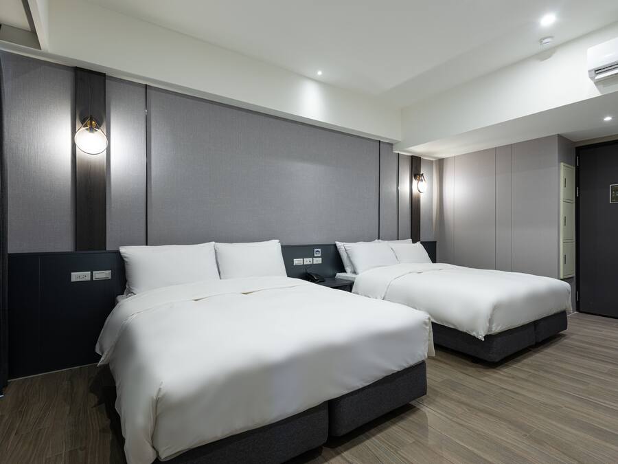 A hotel room with two beds, a dark-colored headboard, a wall-mounted light, and a control panel on the wall.
