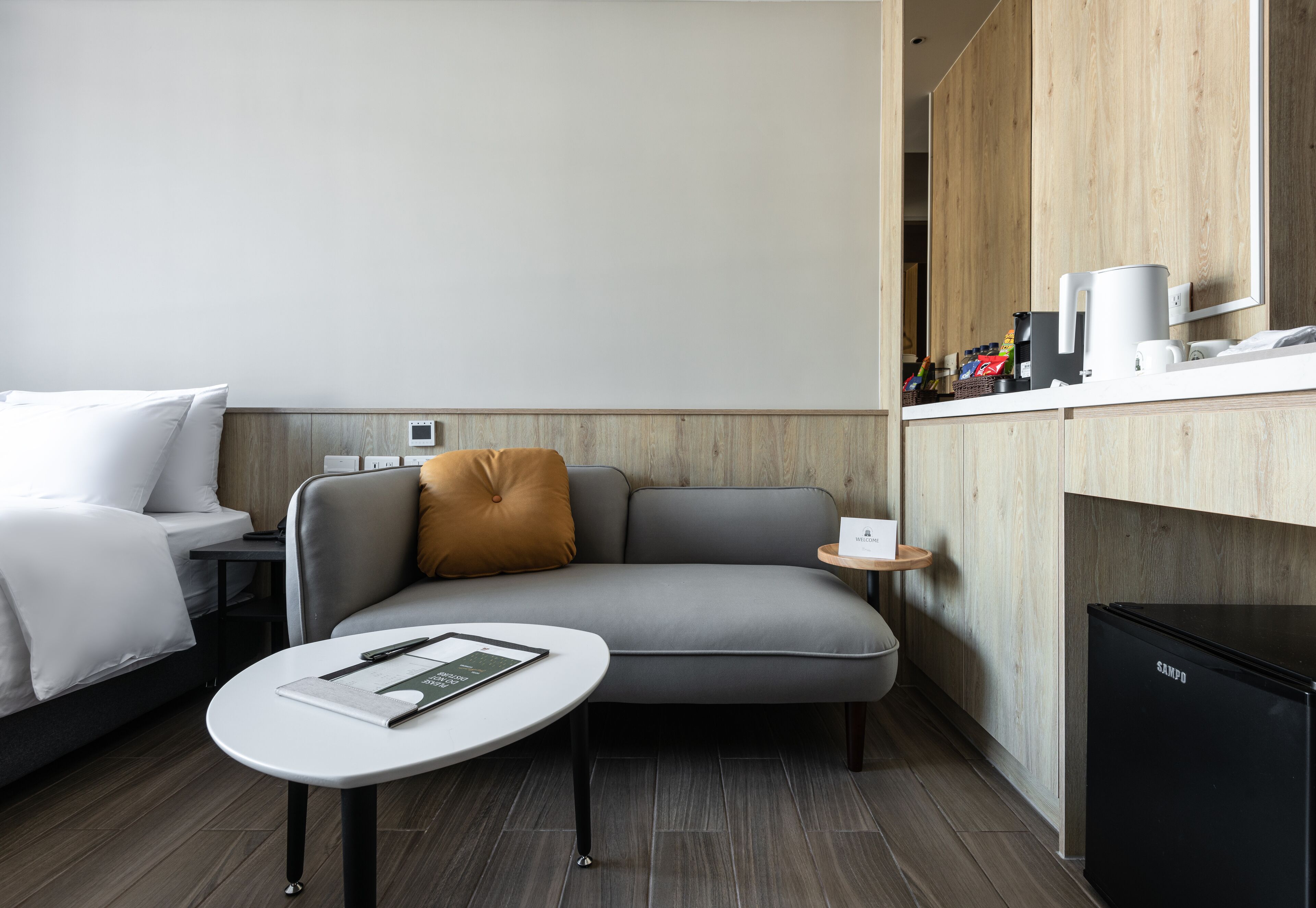 A modern hotel room with a grey sofa, a small round table with a book, a black refrigerator, and a wooden cabinet with a coffee maker.