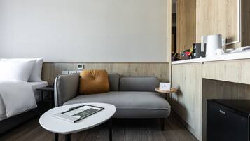 Una habitación de hotel moderna con un sofá gris, una pequeña mesa redonda con un libro, un refrigerador negro y un mueble de madera con una cafetera.
