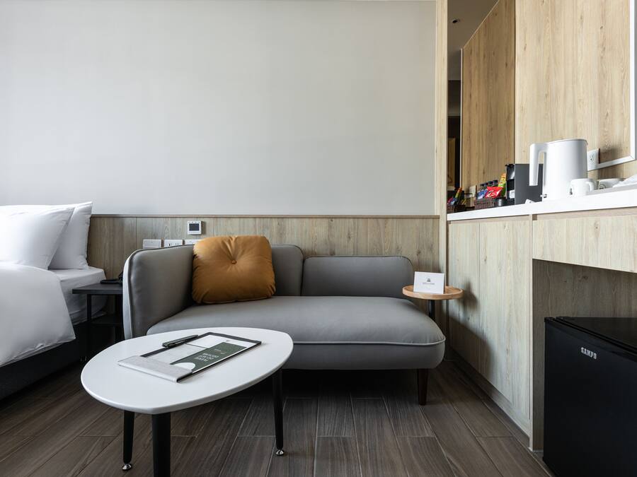 Una habitación de hotel moderna con un sofá gris, una pequeña mesa redonda con un libro, un refrigerador negro y un mueble de madera con una cafetera.